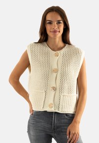 Gilet en maille de couleur crème avec de grandes poches frontales, présentant un motif texturé, des boutons ronds et un design décontracté sans manches.