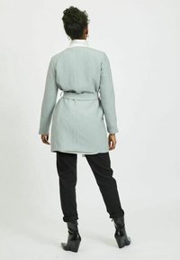 Giacca grigia su misura con cintura in vita, maniche lunghe, spacco posteriore e sotto giacca bianco. Abbinata a pantaloni neri e stivaletti.