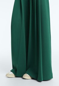 Lange, dunkelgrüne Hosen mit weitem Bein, die teilweise beige und weiße Schuhe auf einem schlichten hellen Hintergrund bedecken.