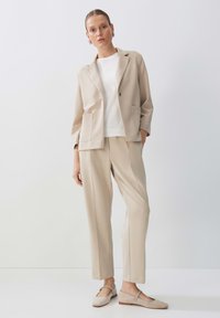 Completo beige con giacca su misura e pantaloni affusolati, abbinato a un top bianco. Tessuto strutturato, tasche laterali e scarpe semplici flat.