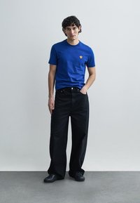 Zila kokvilnas T-krekls ar mazu dzeltenu logotipu uz krūtīm, kombineēts ar brīvi pieguļošām melnām džinsa biksēm un melniem sporta apaviem.