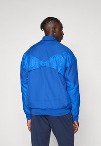 Modrá bunda na zips s textúrovaným chrbtom a rukávmi s jemným vzorom. Mäkká tkanina, vysoký golier a elastický spodný okraj.