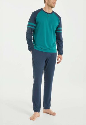 Uomo in piedi a piedi nudi che indossa una maglia a maniche lunghe verde acqua con maniche e strisce blu navy, e pantaloni blu navy su sfondo semplice.