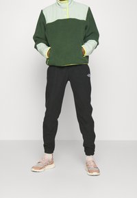 Zielony polar z jasnozieloną górną częścią, żółtymi akcentami, czarnymi spodniami dresowymi i beżowymi butami sportowymi. Teksturowany materiał.