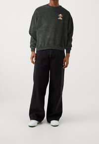 Za duża ciemnoszara bluza z haftowanym wzorem oraz czarne szerokie spodnie typu jeans. Białe sportowe buty z zielonymi akcentami dodają kontrastu stylizacji.