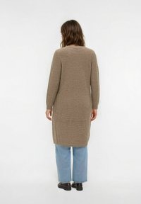 Langer, gerippter, taupefarbener Cardigan mit langen Ärmeln, Seitenschlitzen und lockerer Passform, kombiniert mit hellblauen, weit geschnittenen Jeans und schwarzen Schuhen.