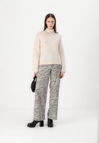 Hellbeige Rollkragenpullover mit weicher Textur; weite graue Hosen mit Zebramuster; schwarze Plateau-Stiefel; hält eine kleine schwarze Handtasche.