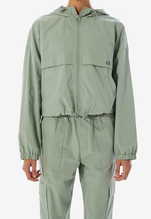 Persoon in een lichtgroene capuchonvest met rits en bijpassende broek met elastische boorden en een taillekoord, staand tegen een witte achtergrond.