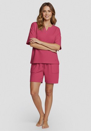 Kobieta stoi boso ubrana w pasujący, luźny różowy top z krótkim rękawem oraz szorty ze sznurkiem, uśmiechając się ze skrzyżowanymi rękami na jednolitym tle.