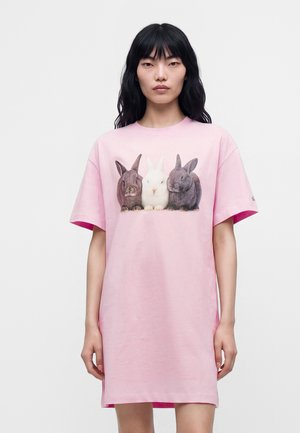Femme portant un t-shirt rose oversize avec trois lapins imprimés sur le devant, debout devant un fond blanc uni.