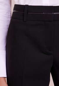 Pantalon noir au toucher lisse, avec une coupe ajustée, des passants de ceinture et des détails de couture subtils. Associé à une chemise blanche à manches longues.