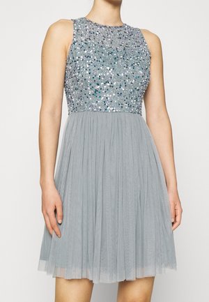 Vestido sin mangas gris claro con cuerpo de lentejuelas y falda de tul plisada, llevado por una persona de pie con los brazos relajados a los lados.