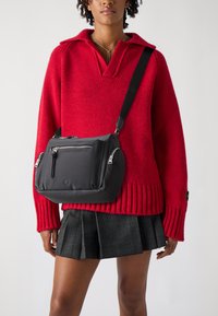 Borsa a tracolla nera con multiple tasche con cerniera, materiale liscio, forma arrotondata e tracolla regolabile, indossata sopra un maglione rosso.