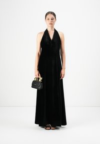 Woman wearing a black velvet halter-neck gown, holding a small black handbag with gold handles, wearing black strappy sandals.