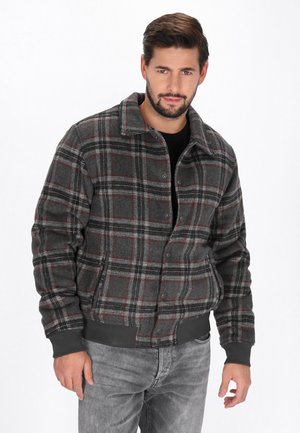 Bearded man wearing a gray plaid button-up jacket with black cuffs and collar, standing with one hand relaxed by his side.
