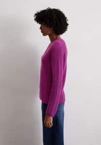 Fuchsia Strickpulli mit tiefem V-Ausschnitt, langen Ärmeln und geripptem Muster. Kombiniert mit dunkelblauen Jeans, von der Seite präsentiert.
