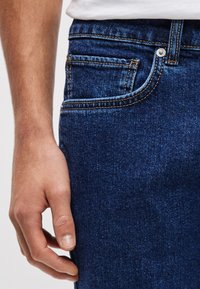 Tumesinised teksapüksid on klassikalise viie tasku disainiga, kontrastse õmbluse ja metallnupuga kinnitusega.