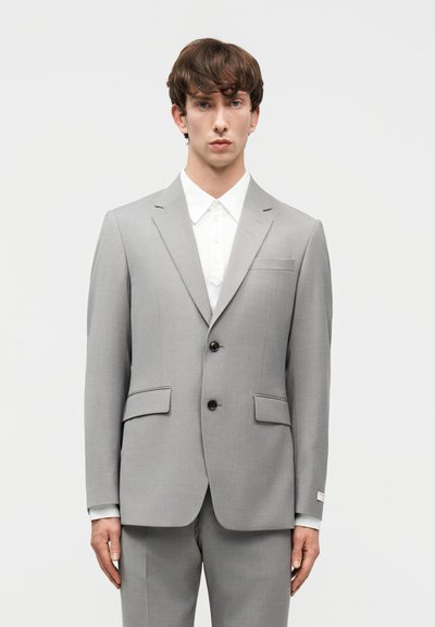 Νεαρός άνδρας στέκεται φορώντας ένα ανοιχτό γκρι σακάκι με δύο κουμπιά πάνω από ένα λευκό πουκάμισο με γιακά, μπροστά σε ένα απλό φωτεινό φόντο.