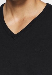 Maglione nero a collo a V in maglia con scollo e polsini a coste, con una discreta firma nella zona del petto. Texture liscia e forma aderente.