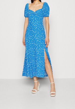 Vrouw draagt een blauwe midi-jurk met witte bloemenprint, pofmouwen, sweetheart halslijn, zijsplit en witte enkellaarsjes met blokhak en enkelbandje.
