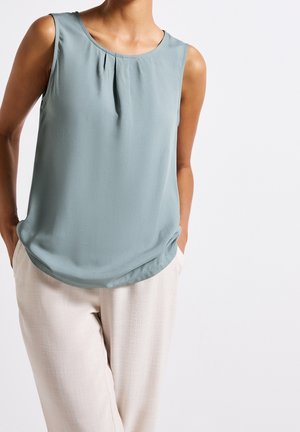 Mouwloze lichtblauwe blouse met plooien bij de halslijn gecombineerd met beige, gestructureerde broek met hoge taille, handen in de zakken.