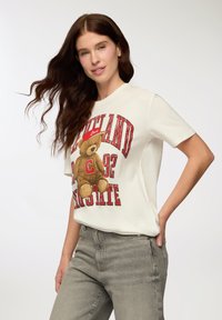 Bawełniany T-shirt w kolorze ecru z grafiką misia i napisem "Cleveland Love" w kolorze czerwonym i czarnym. Krótkie rękawy, luźny krój.