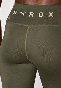Olivgrüne, hochgeschnittene Leggings aus elastischem Stoff, mit Flachnaht und einem kontrastierenden Logo am Bund.