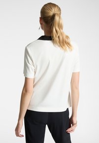 Camicia bianca a maniche corte con colletto nero, caratterizzata da una texture liscia e un design semplice; vista dal retro.