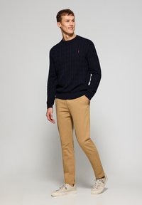 Marineblaue Kabelstrickpullover kombiniert mit beigen Chinos und hellen Sneakers. Der Pullover hat einen Rundhalsausschnitt und gerippte Bündchen.