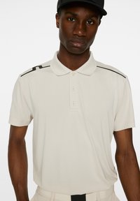 Ljust beige polo med krage, knappslå och svarta axeldetaljer. Slätt, lätt material; korta ärmar.