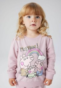 Noor blonde lokkis, kellel on lokkis juuksed, kannab roosat sviitrit, millel on koomiksikad, vikerkaar ja tekst "Mänguaeg!" heledal taustal.