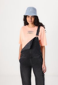 Oranje t-shirt met een grafisch ontwerp in combinatie met zwarte overalls. Een lichtblauwe emmerhoed maakt de outfit compleet. De overalls hebben verstelbare banden.