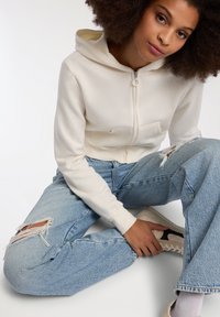 Kremowa, krótka bluza z kapturem na zamek, uszyta z miękkiego materiału, w połączeniu z luźnymi jasnoniebieskimi, przetartymi dżinsami. Białe sneakersy dopełniają całość.