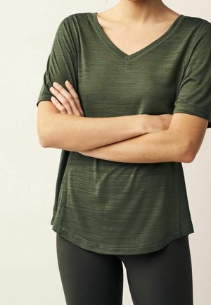 Persoon draagt een olijfgroen T-shirt met korte mouwen en V-hals en zwarte leggings, staand met gekruiste armen tegen een lichte achtergrond.