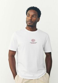Hvid kortærmet t-shirt med et rødt blomsterdesign og teksten "ASAHIKAWA Hokkaido" på brystet. Blødt bomuldsstof, afslappet pasform.