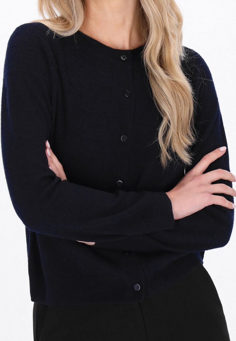 Femme portant un cardigan noir boutonné, cheveux longs et blonds, les bras croisés sur la poitrine, sur fond blanc.