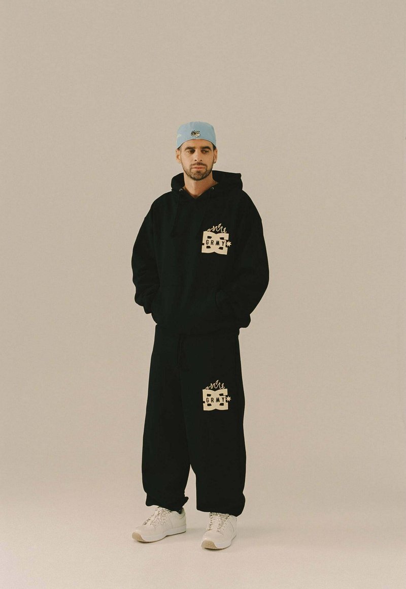 Man in een zwarte hoodie en broek met bijpassende vlamlogo's, witte sneakers en een lichtblauwe pet, staande tegen een effen achtergrond.