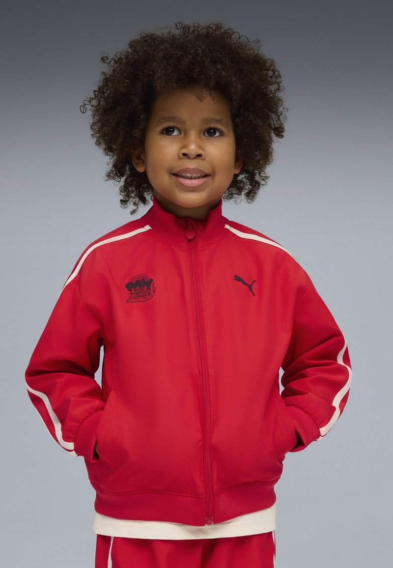 Jeune enfant aux cheveux bouclés portant un survêtement rouge Puma, les mains dans les poches, souriant et regardant légèrement vers la gauche sur un fond uni.