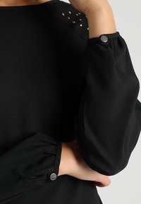 Chemisier noir avec des manches bouffantes et des poignets boutonnés. Présente des clous métalliques sur les épaules, une texture lisse et un design décontracté.