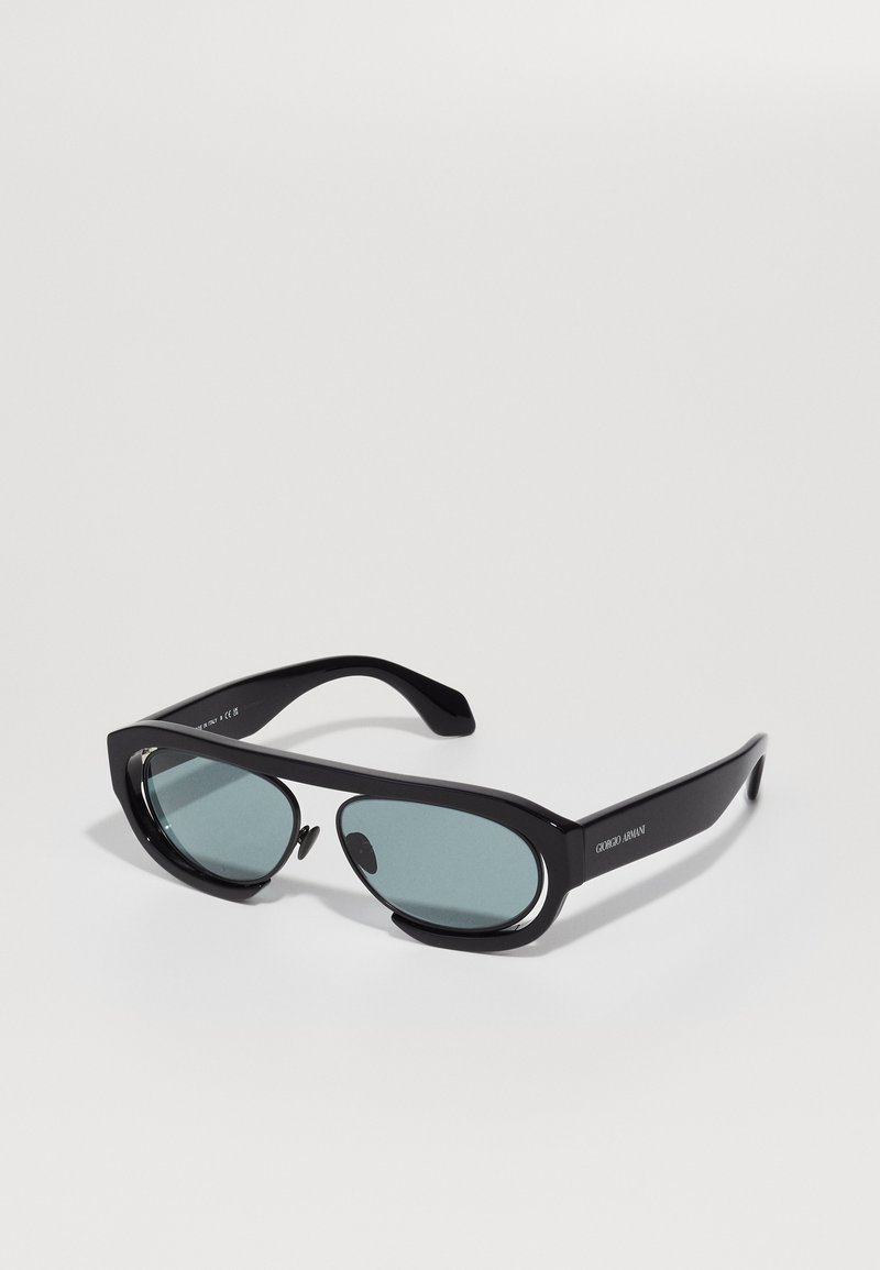 Ochelari de soare negri, eleganți, cu lentile verzi, având o formă distinctivă și un logo pe braț, așezați pe un fundal alb simplu.