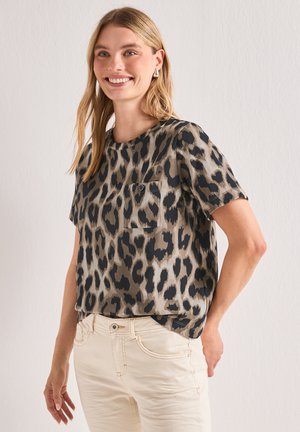 Smilende kvinde iført en kortærmet leopardmønstret top og lyse beige bukser, stående med den ene hånd bag ryggen.