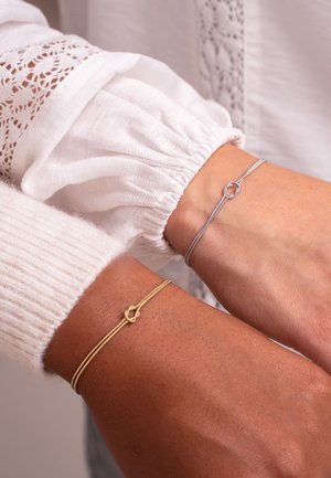 Due braccialetti sui polsi: uno dorato con un dettaglio a nodo, l'altro argentato, entrambi con design sottili ed eleganti. Maniche bianche con texture visibili.