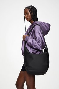 Črna ramenska torbica iz gladkega materiala, s preprosto obliko in trakcem, ki jo drži oseba v vijolični jakni.