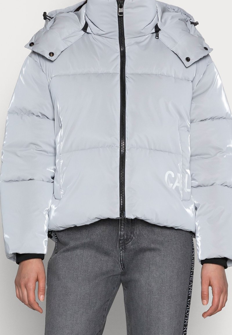 Jasnoszara błyszcząca kurtka puchowa z czarnym zamkiem, odpinanym kapturem i subtelnym logo po prawej dolnej stronie, noszona z szarymi dżinsami.