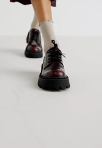 Scarpe di pelle borgogna con suole nere spesse, punte rotonde e lacci neri; indossate con calzini a coste di un leggero beige.
