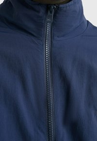 Mörkblå zip-up jacka med hög krage, som har en vattentät yta och ett centralt blixtlås. Slät tyg, minimalistisk design.