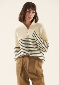 Krēmkrāsas ribota džemperis ar melnām horizontālām svītrām un pusrāvēju apkakli, kopā ar gaiši brūnām plisētām biksēm.