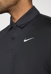 Polo noir avec un col boutonné, fabriqué en tissu lisse. Présente un logo Nike blanc sur la poitrine gauche et des détails de couture subtils.
