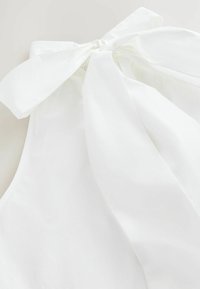 Top bianco con collo all'americana realizzato in tessuto leggero, caratterizzato da un grande fiocco sul retro e una superficie liscia.