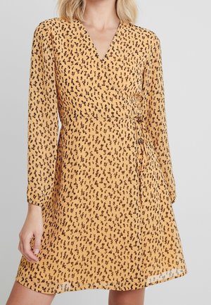 Woman wearing a yellow wrap dress with long sleeves and small brown leaf patterns on a plain background.